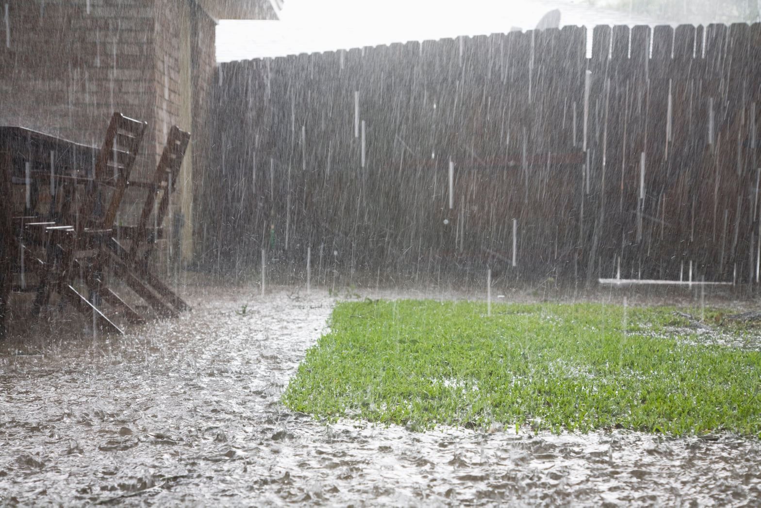 Rain falling heavily in a backyard with a green lawn, wooden fence, and chairs.