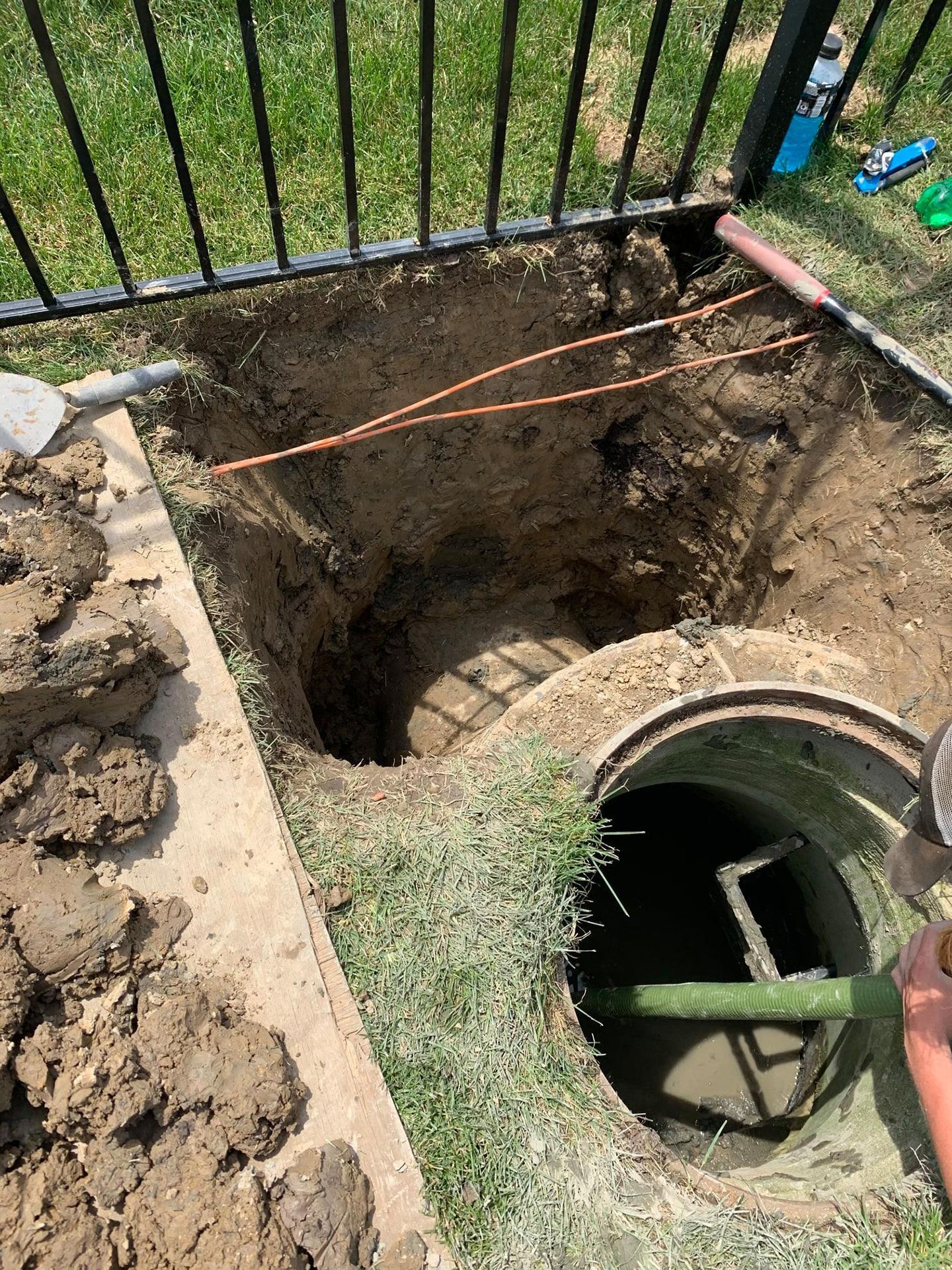 A deep hole next to a metal fence exposes a septic tank; dirt and tools are visible.