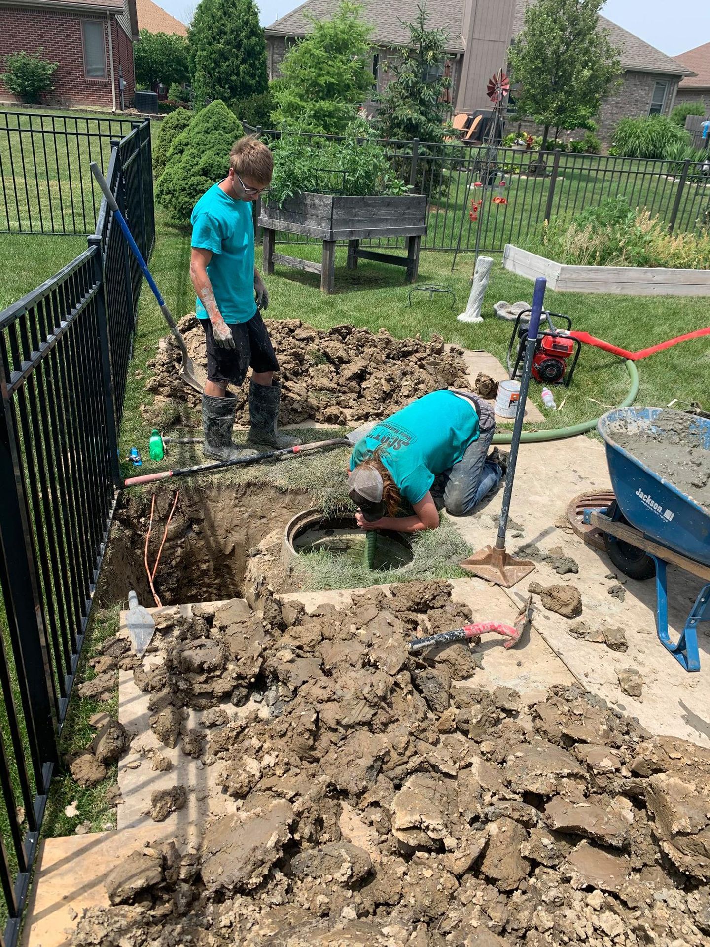 Two people digging near a fence, dirt pile, and cement mixer on a sunny day.