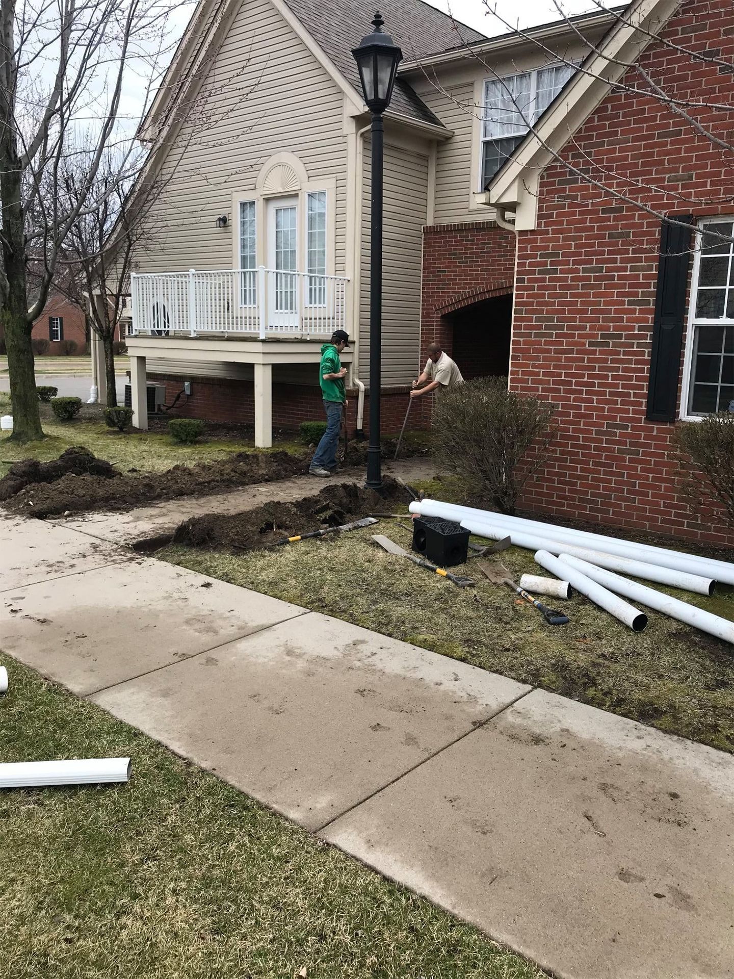 People installing irrigation pipes in front of a two-story house with a brick facade and a light pole.
