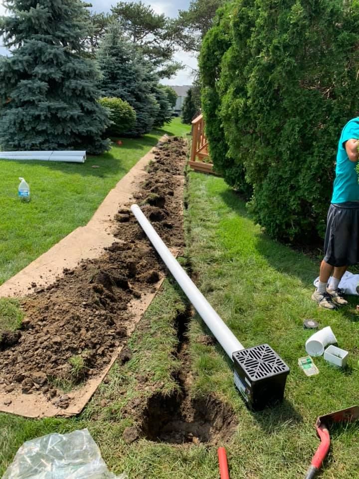 A trench dug in a lawn, with a drain and pipe laid out, near a person standing.