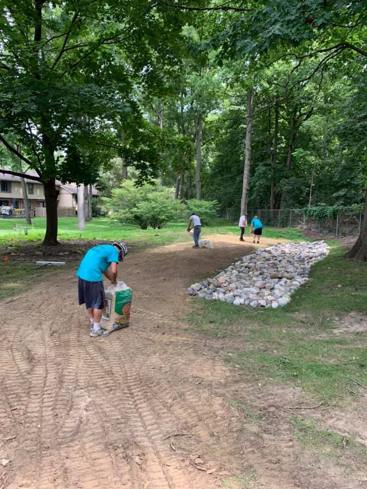 Workers installing landscaping in a wooded area; one man holds a bag, three others stand nearby.