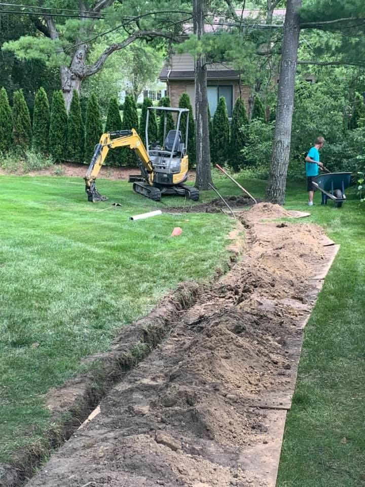 A mini excavator digs a trench in a grassy backyard, a person works with a wheelbarrow nearby.