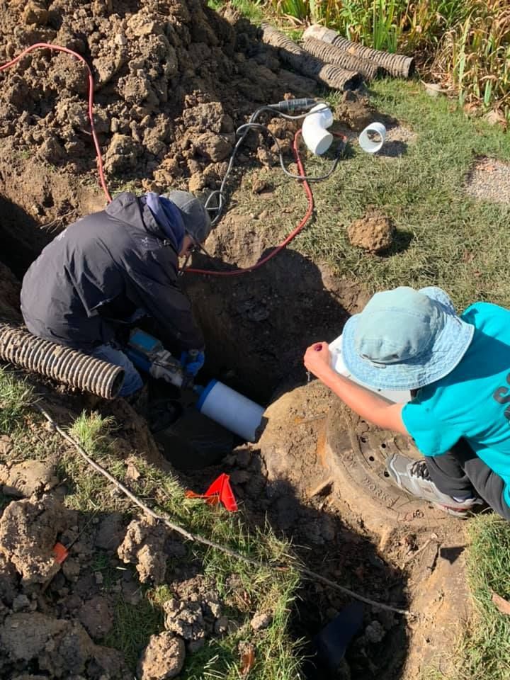Two people working on plumbing in a trench. One uses a tool, the other observes. Outdoors, daylight.