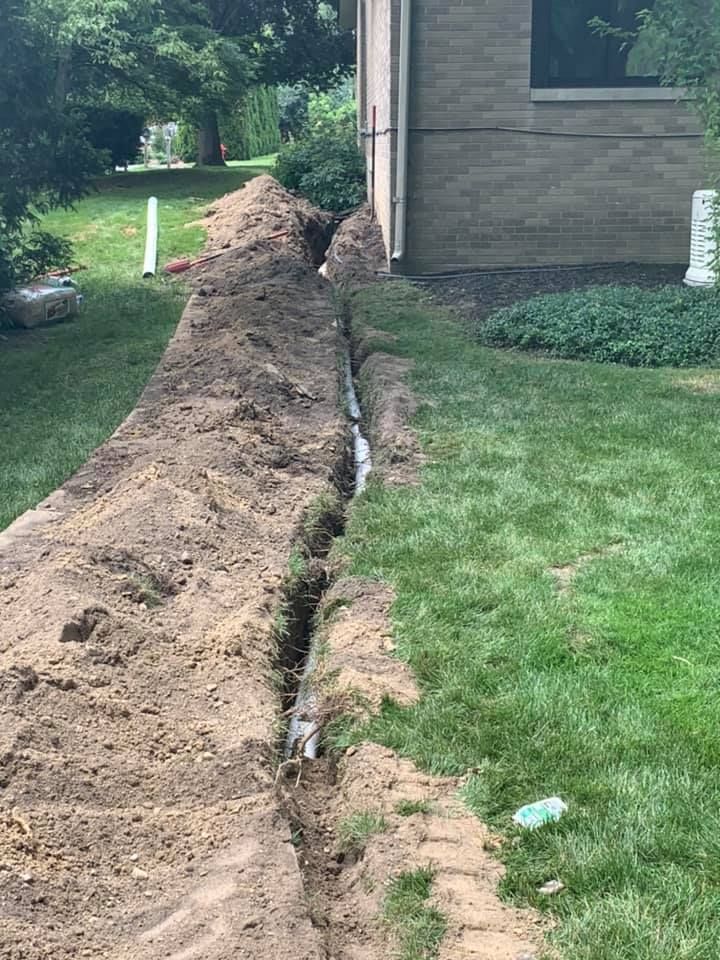A long trench dug alongside a building, likely for drainage or utility lines, on a grassy lawn.