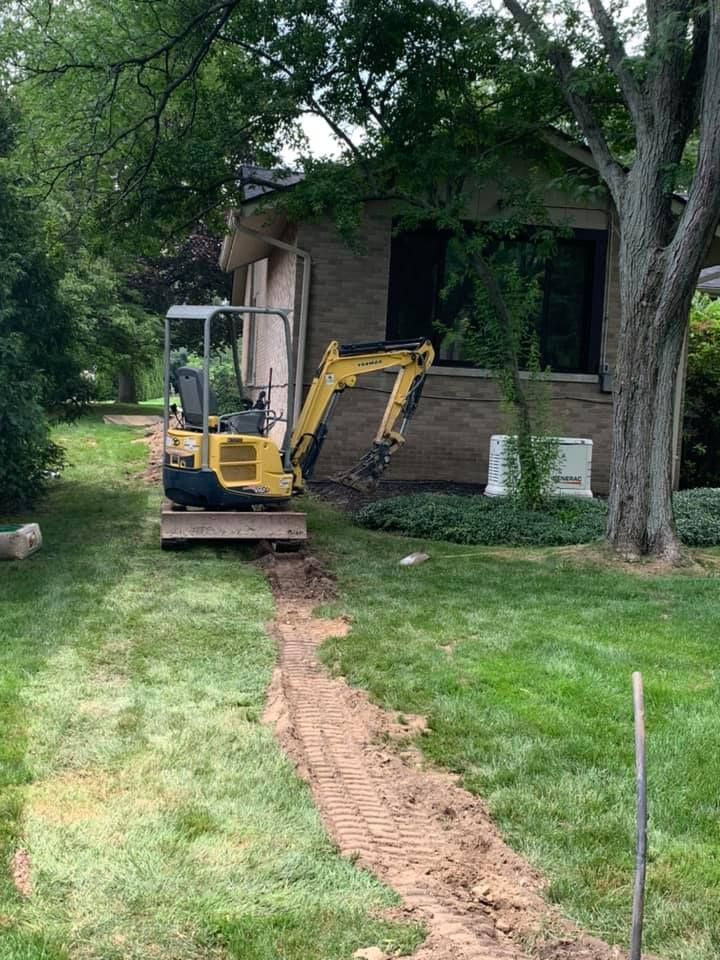 A small yellow excavator on a lawn next to a building, digging a trench in the grass.