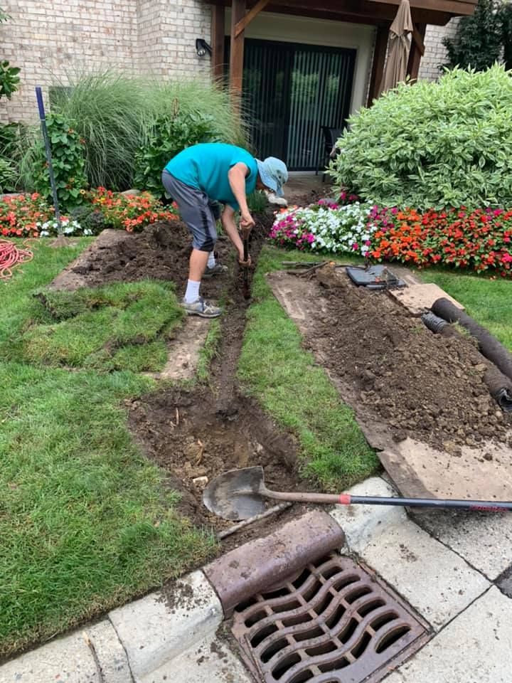 Person digging a trench in a yard near flowers and a drain. They are wearing a hat, shorts, and using a shovel.