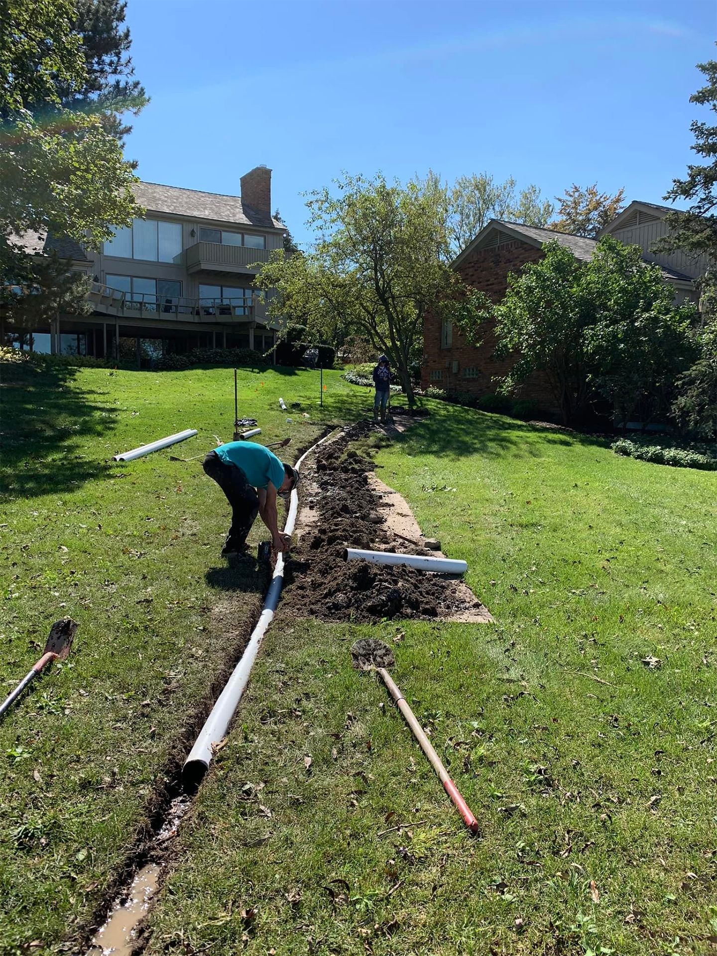 Workers installing drainage pipes in a grassy yard, near a house under a clear sky.