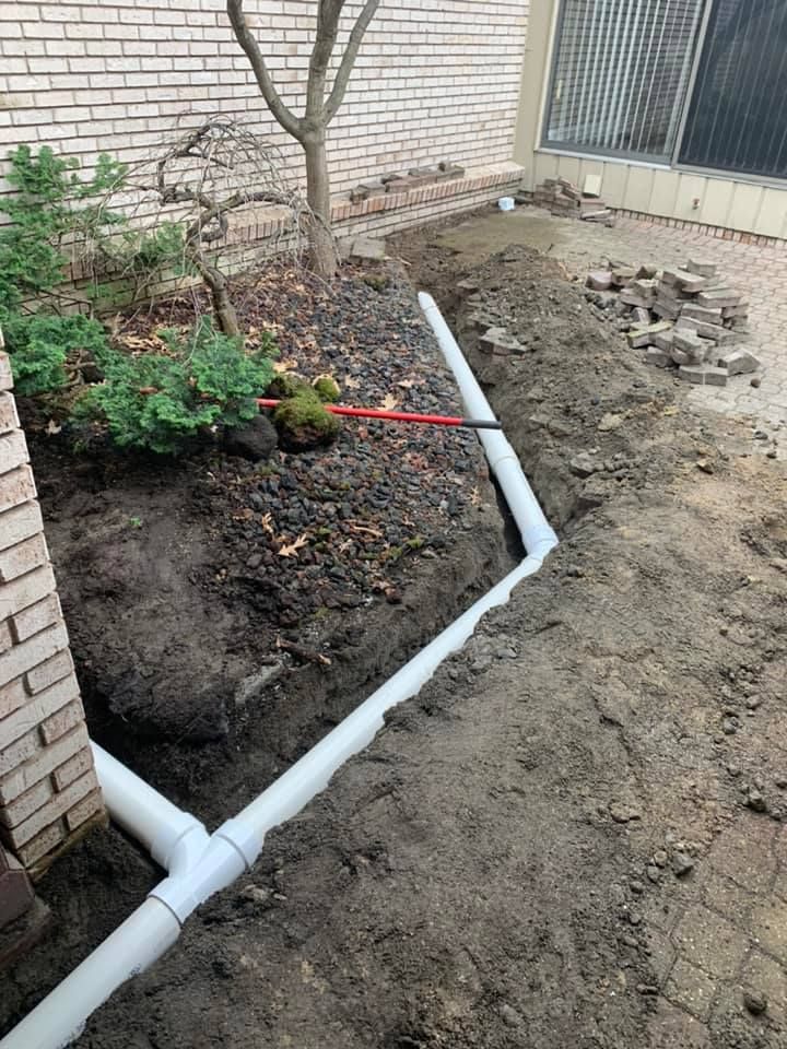 White PVC pipes installed in a trench along a brick wall and a garden bed. Soil and small plants are present.