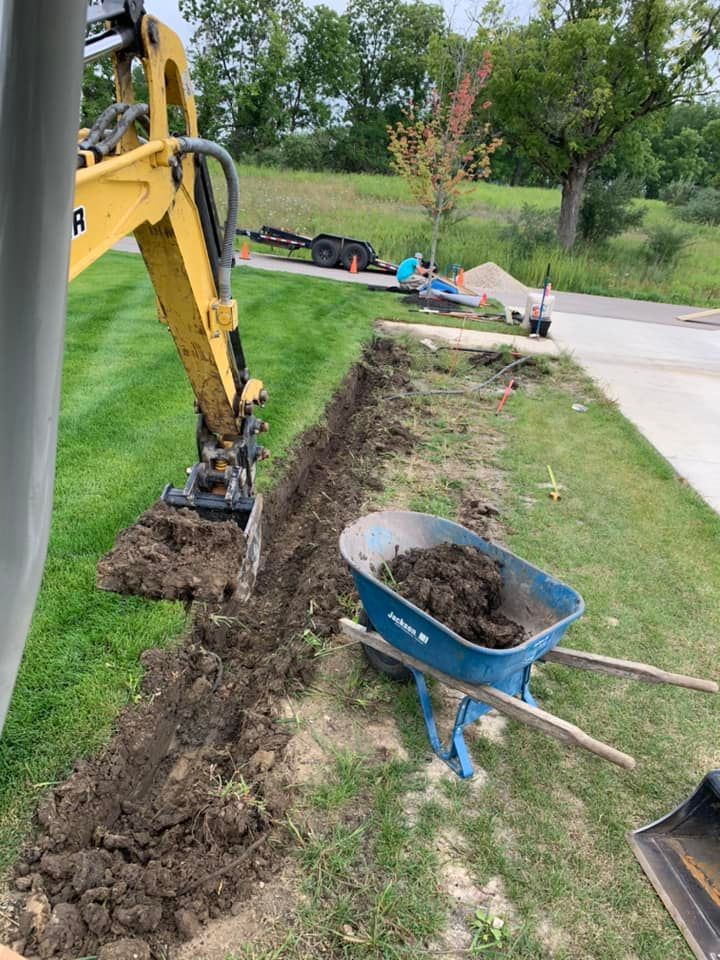 Yellow excavator digging a trench next to a sidewalk and grass. Wheelbarrow full of dirt nearby.