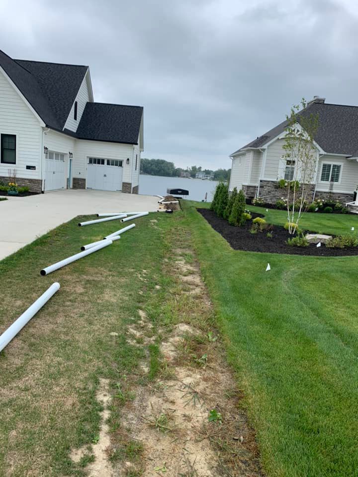 White pipes lie on grass beside a driveway, leading towards two white houses with dark roofs.