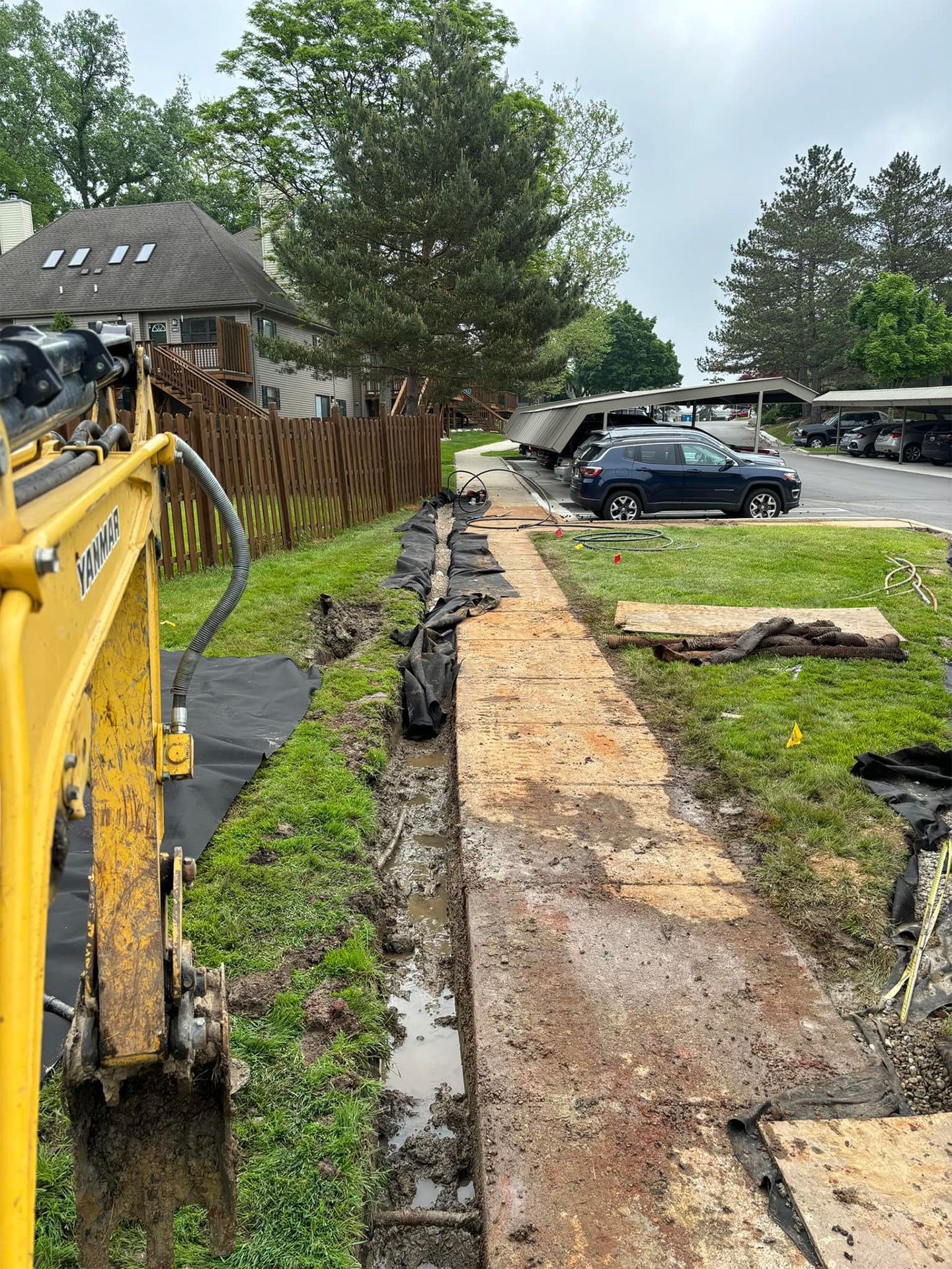 Yellow excavator digging trench along a muddy sidewalk.