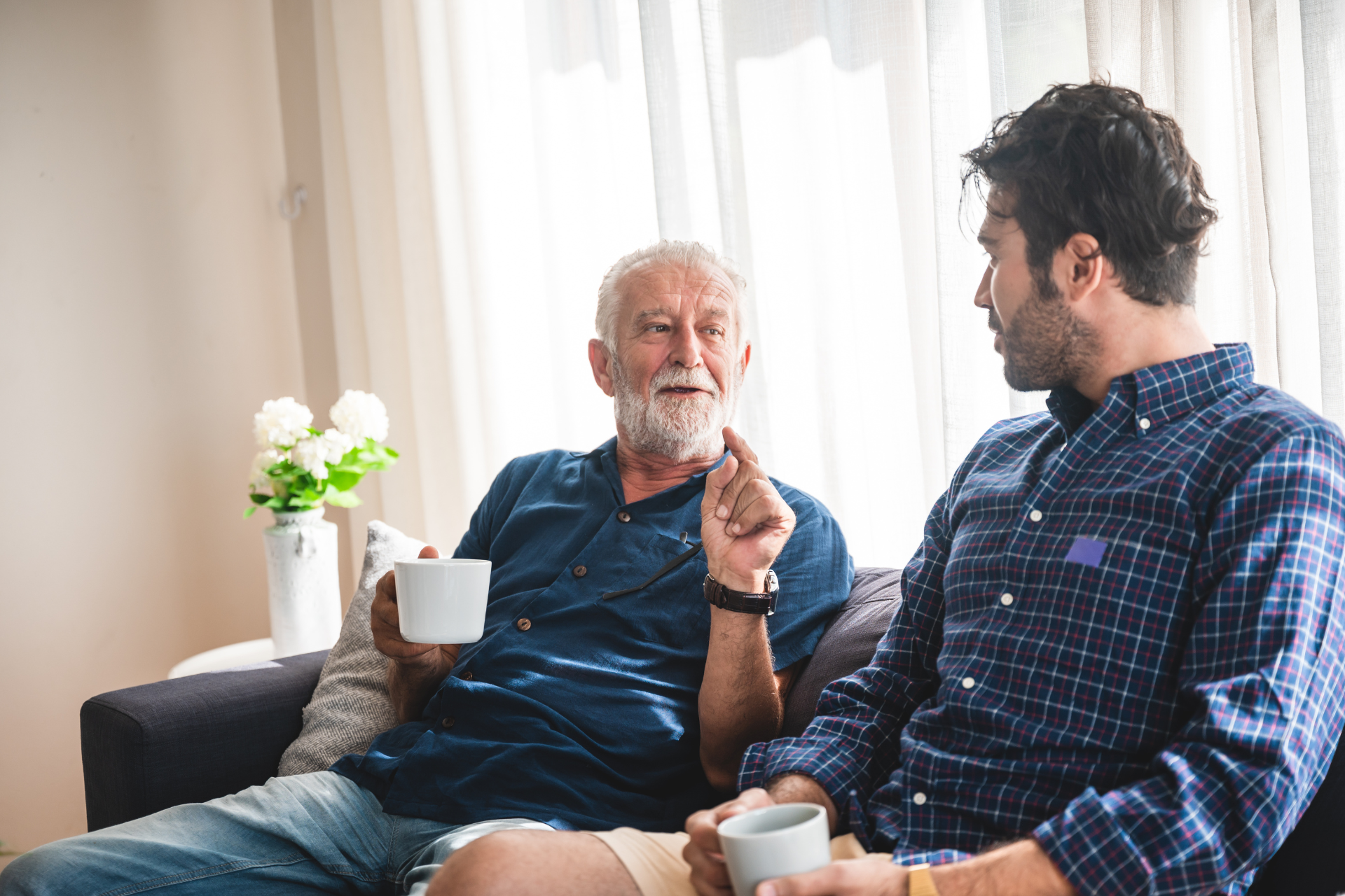 Older man with son sitting on a couch drinking coffee and talking.