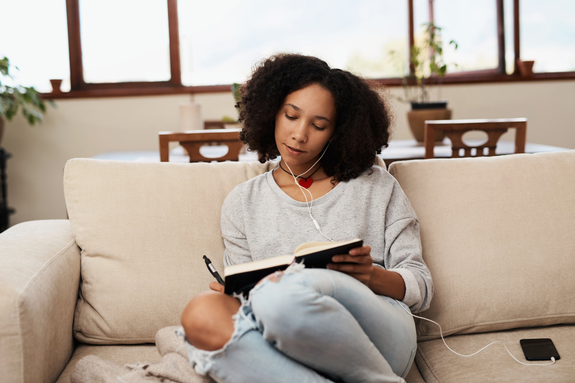 A woman is sitting on a couch reading a book and listening to music.