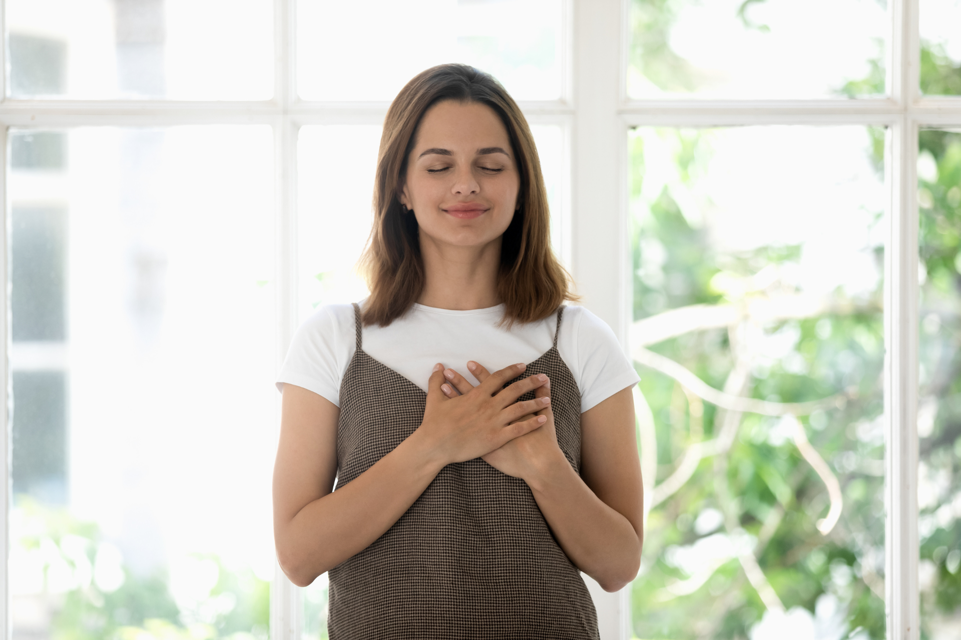 A woman is holding her hands to her chest in front of a window.