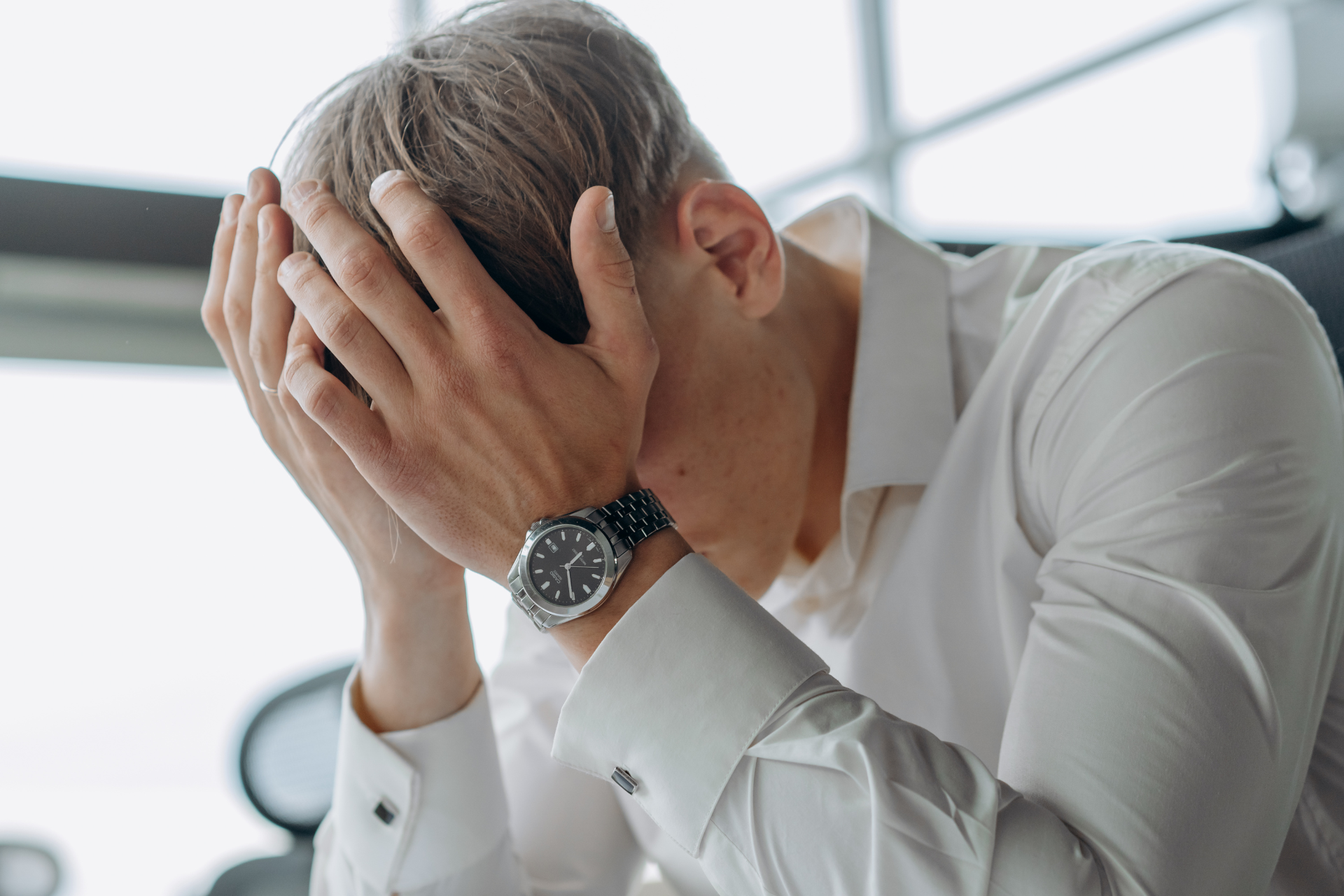 A man in a white shirt is covering his face with his hands.