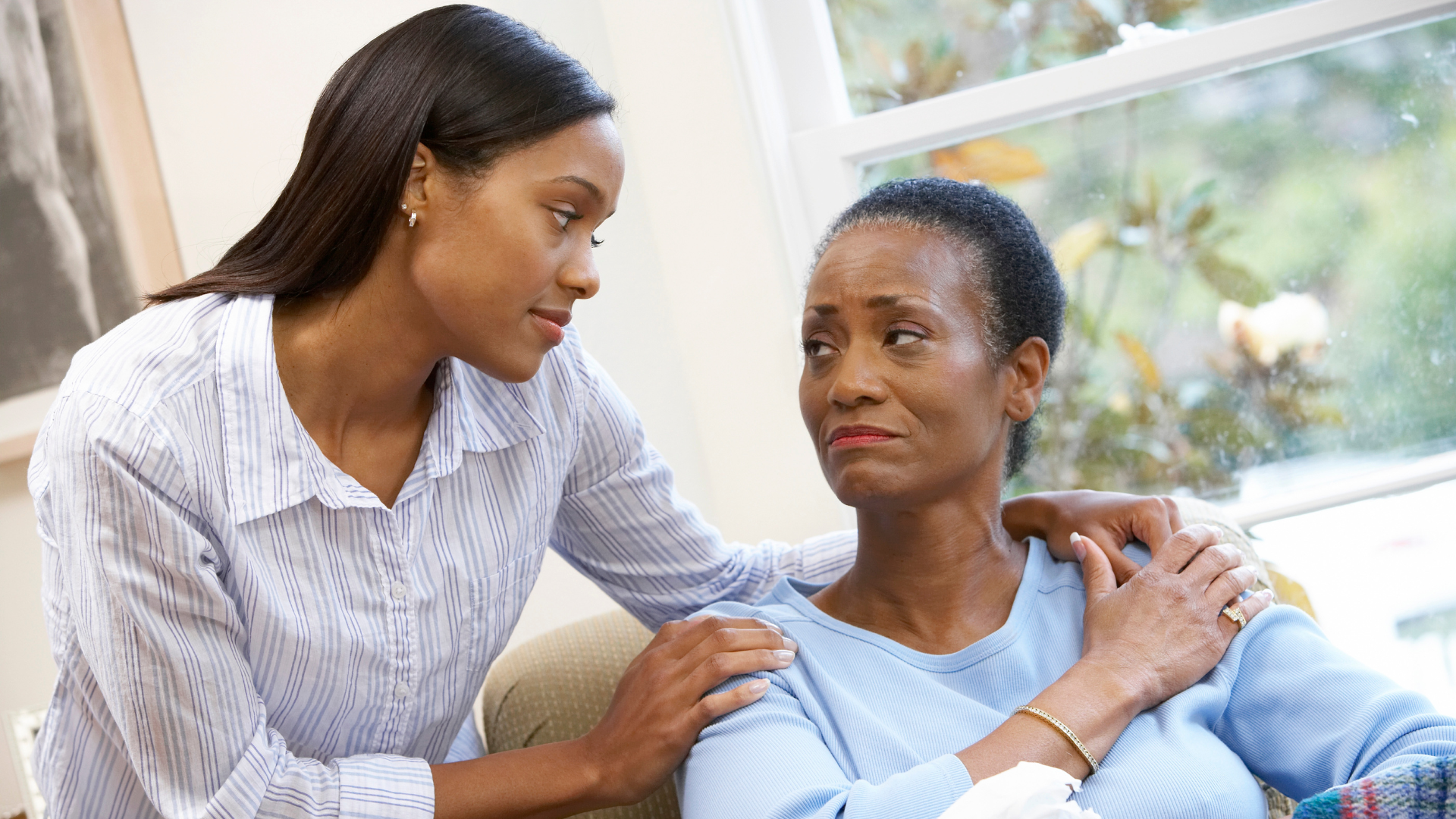 A woman is comforting an older woman who is sitting on a couch.