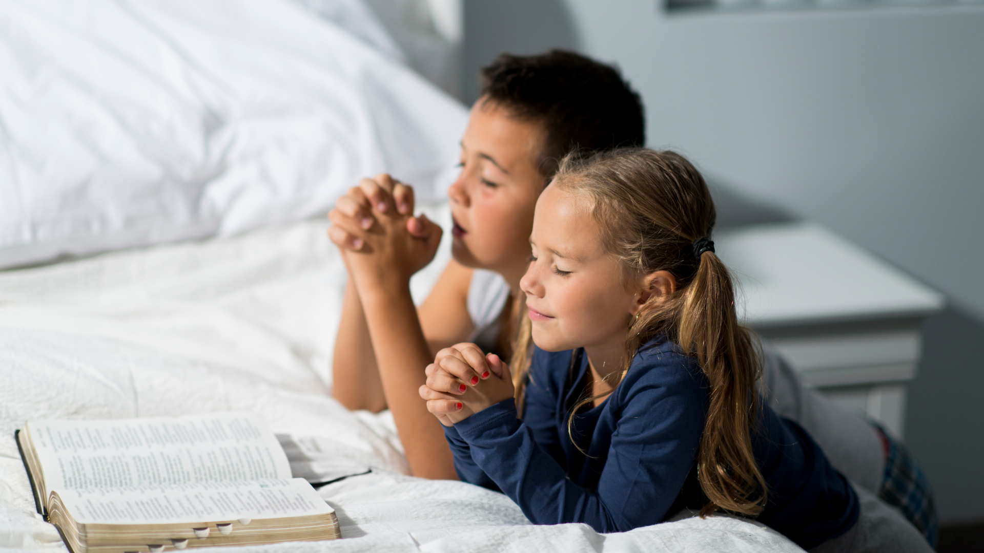 A boy and a girl are praying on a bed with a bible.