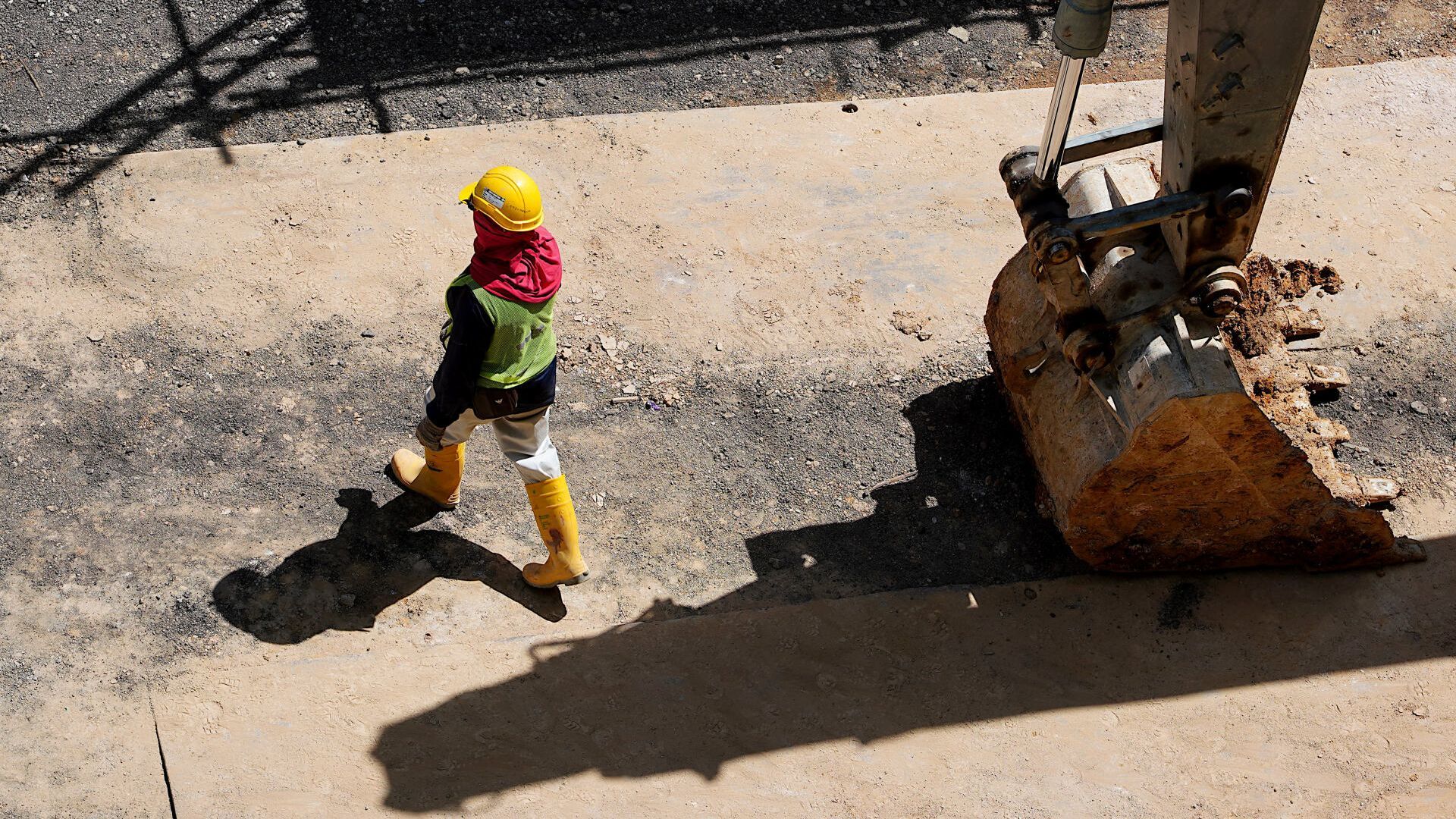 A construction worker wearing a hard hat and yellow boots is walking next to a large excavator bucket.