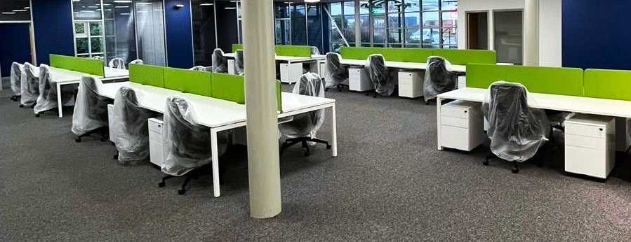 Empty office with rows of white desks, green dividers, and chairs covered in plastic. Gray carpet, blue walls.
