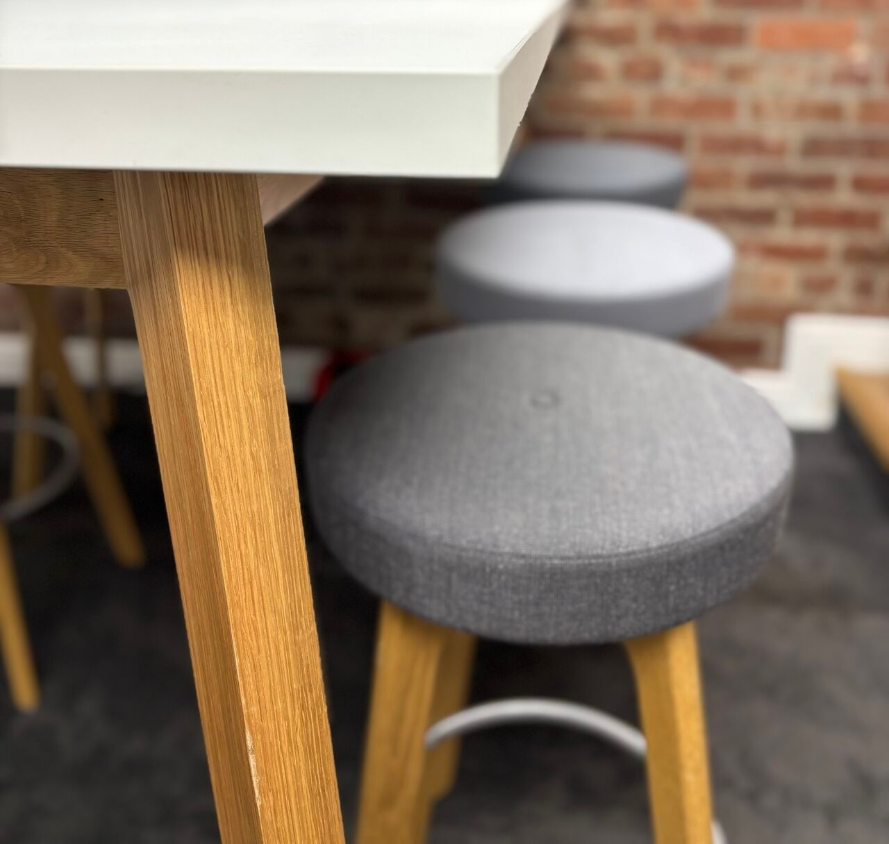 Close-up of a white table edge and a grey padded stool with wooden legs in front of a brick wall.