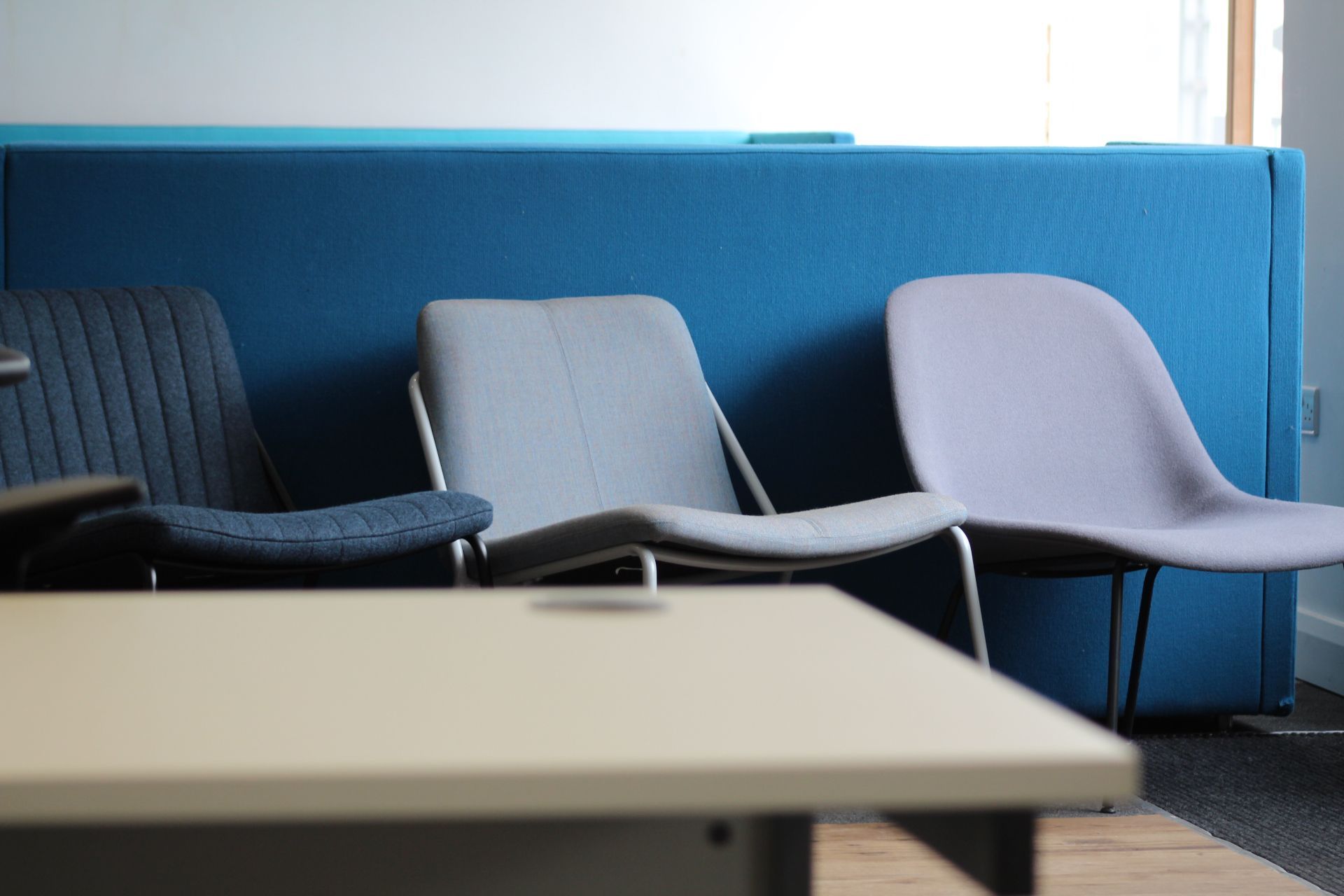 Three chairs in front of a blue wall and a light-colored desk, the chairs are different colors and styles.