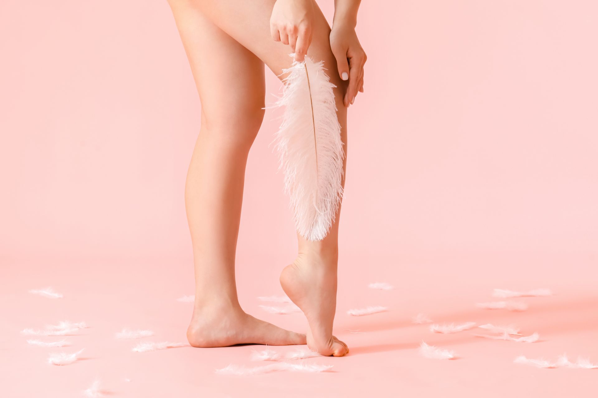 A woman is holding a white feather on her leg on a pink background.