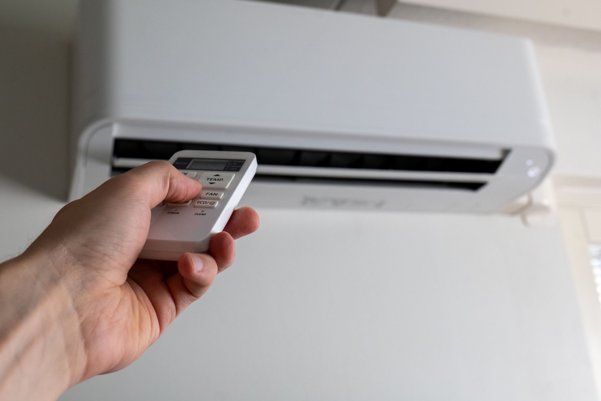 Hand holding a remote control aimed at a wall mounted air conditioner, adjusting cooling settings.