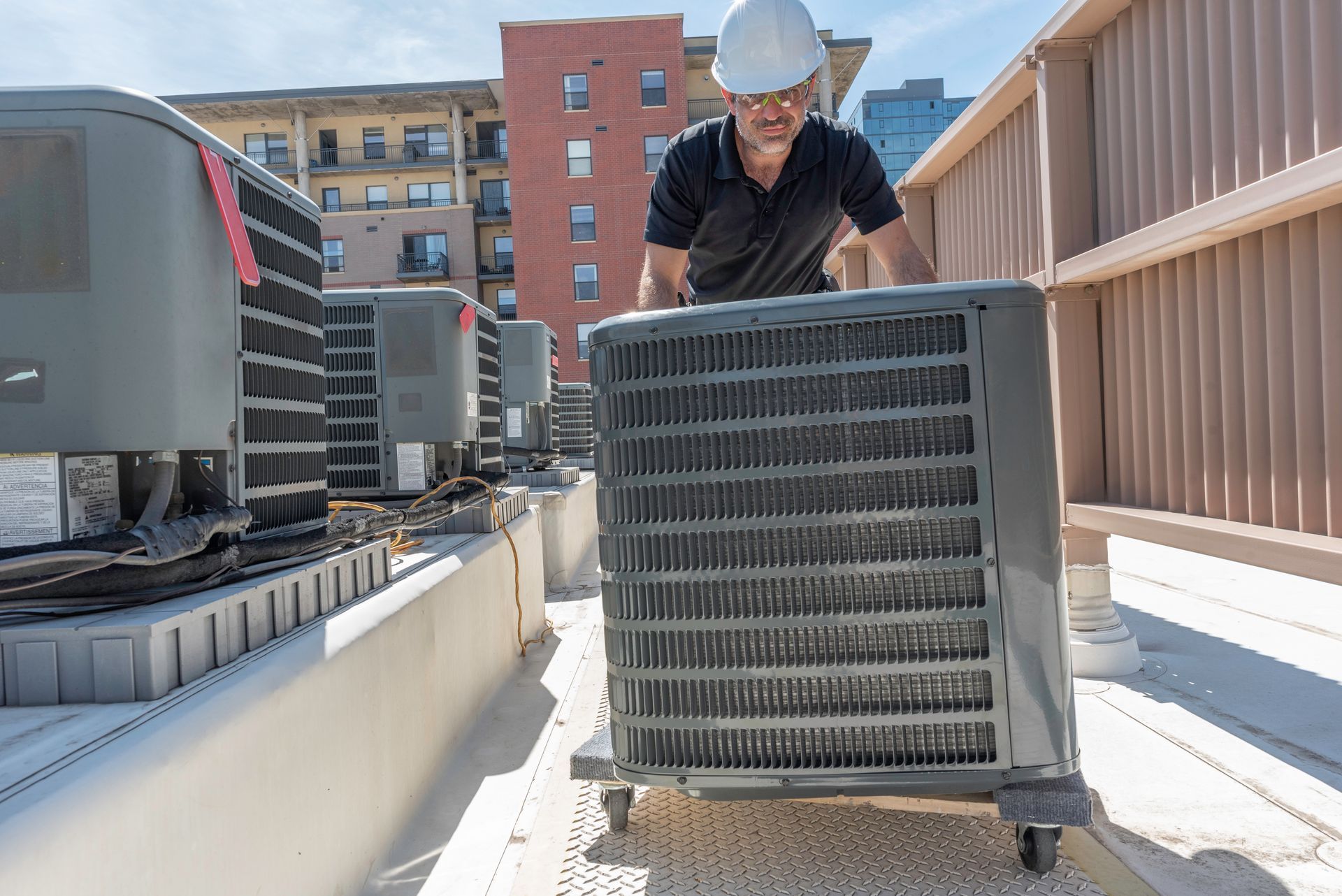 HVAC technician positioning outdoor air conditioning unit on rooftop beside multiple systems.