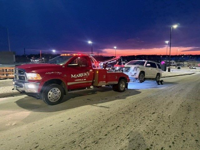 A red Marek's tow truck is towing a white SUV on a road at dusk under a colorful sunset sky.