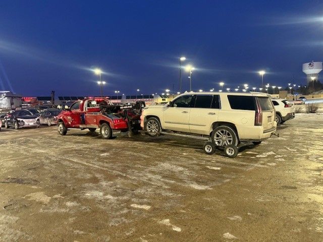 A red tow truck pulling a white Cadillac SUV on a tow dolly in a snowy, dusk-lit parking lot.