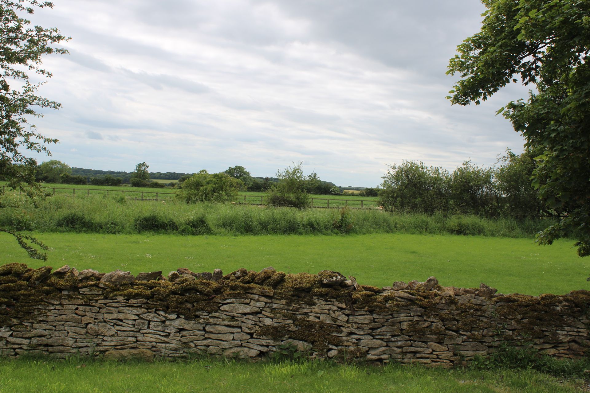 Stone wall beside a green field under a cloudy sky, with trees and hedges in the distance