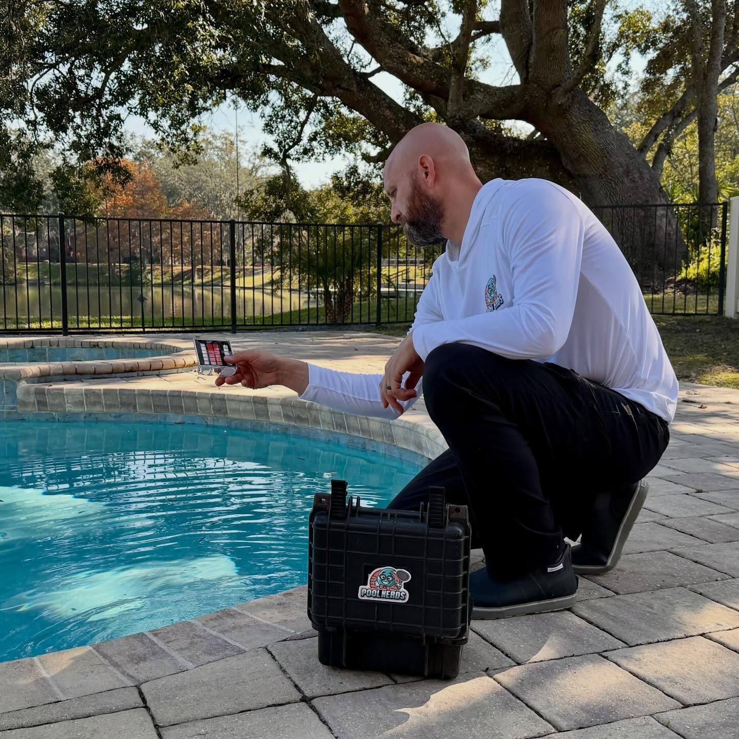 Man squatted by a pool taking a video with a phone. Black case sits nearby. Outdoors on a sunny day.