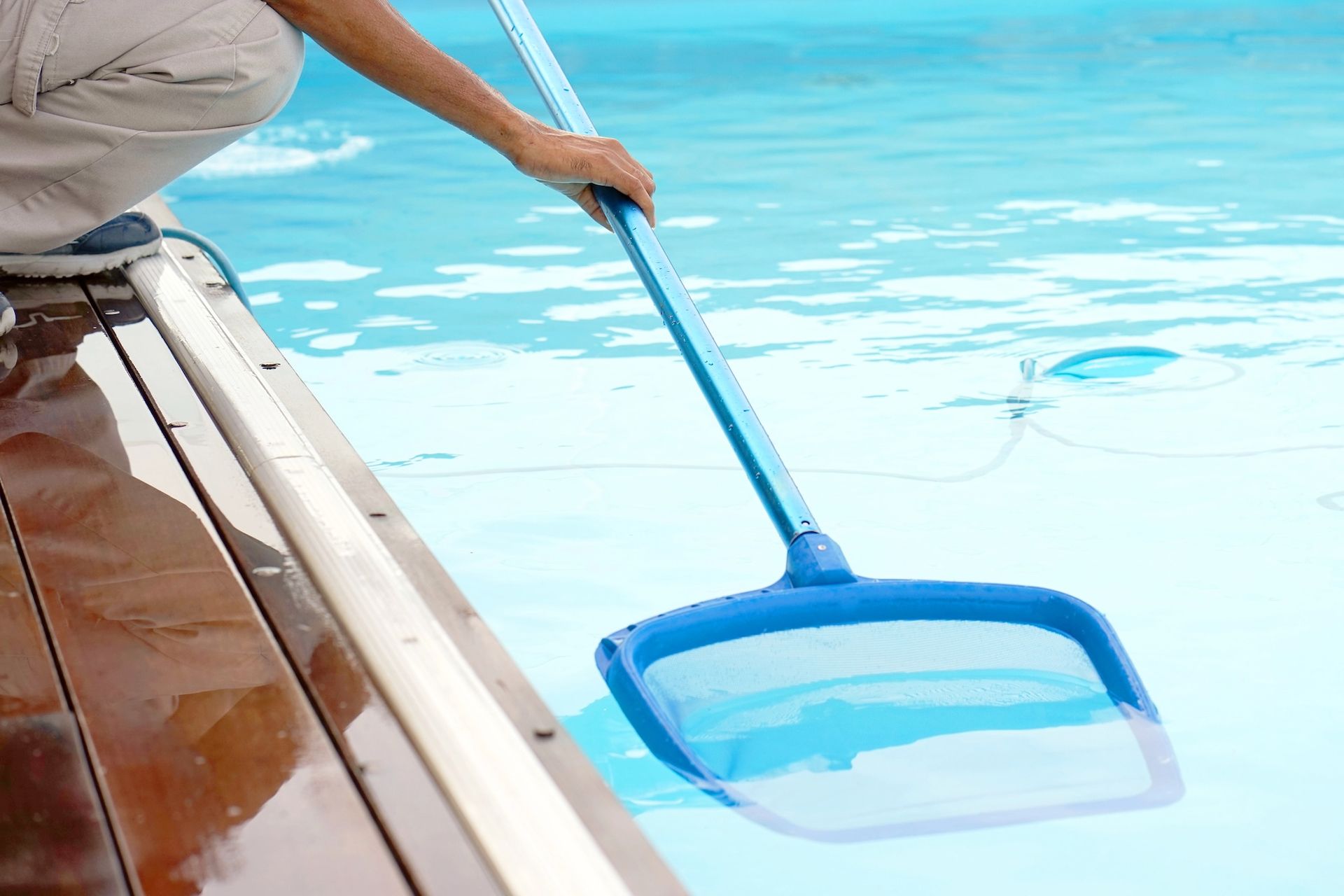 Person using a blue net to scoop debris from a light blue swimming pool.