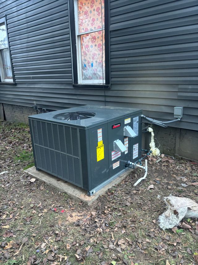 Outdoor HVAC unit beside a dark-sided house with a window and fallen leaves on the ground