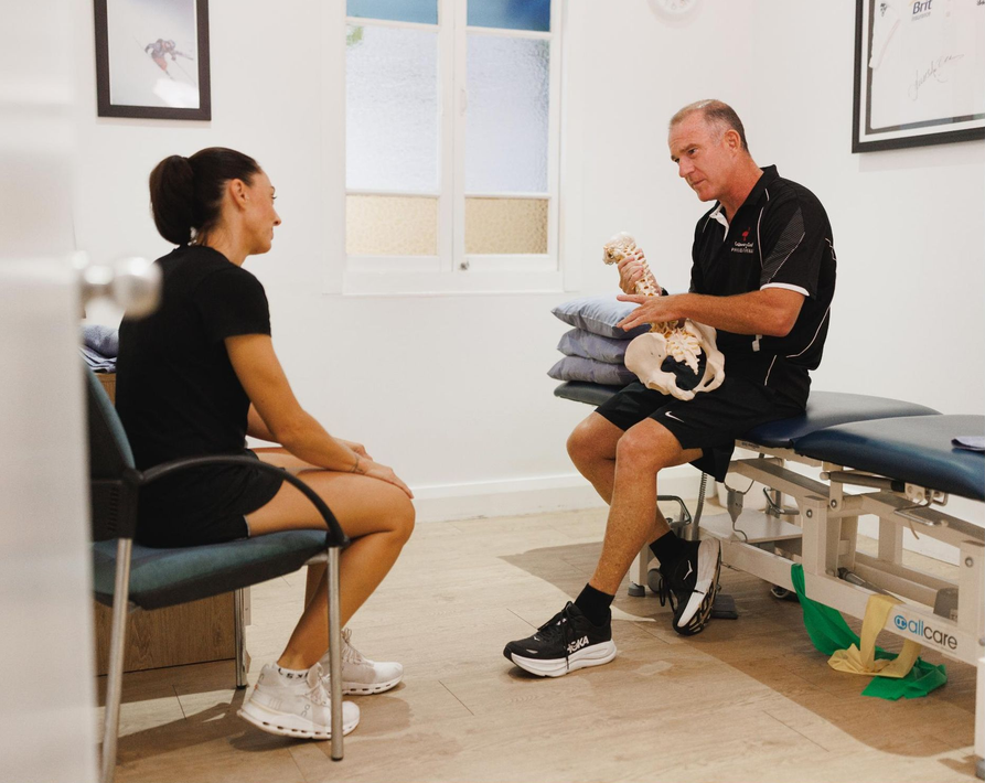 A physical therapist holds a pelvic model to explain an injury to a patient sitting in an office.