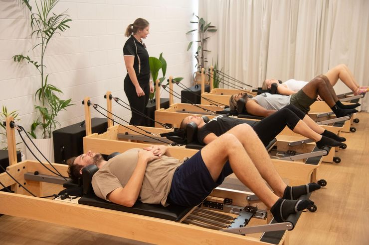 People performing Pilates exercises on reformers in a studio while an instructor watches.