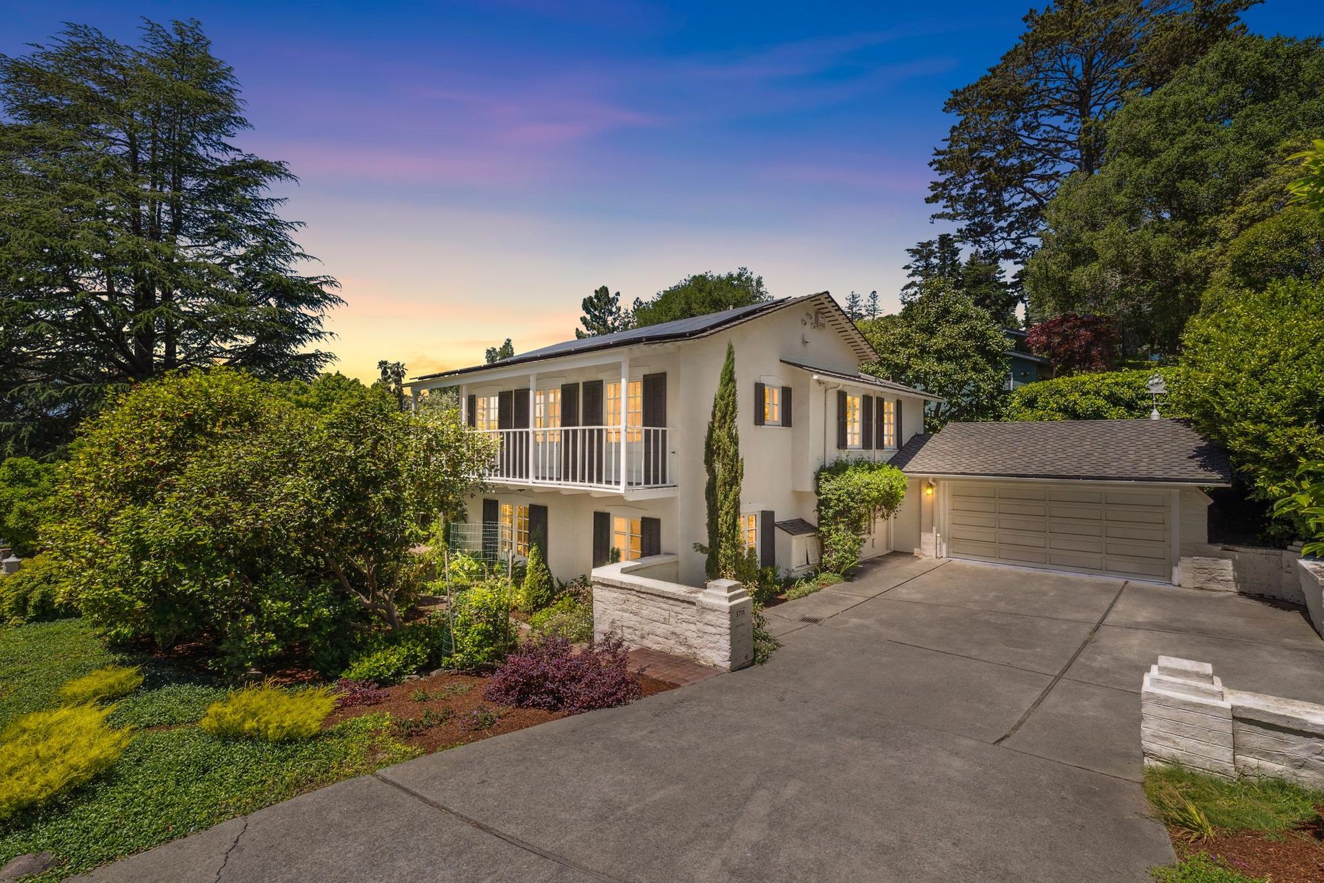 Two-story home with white stucco walls, balcony, and attached garage. Driveway, lush greenery, and sunset sky.