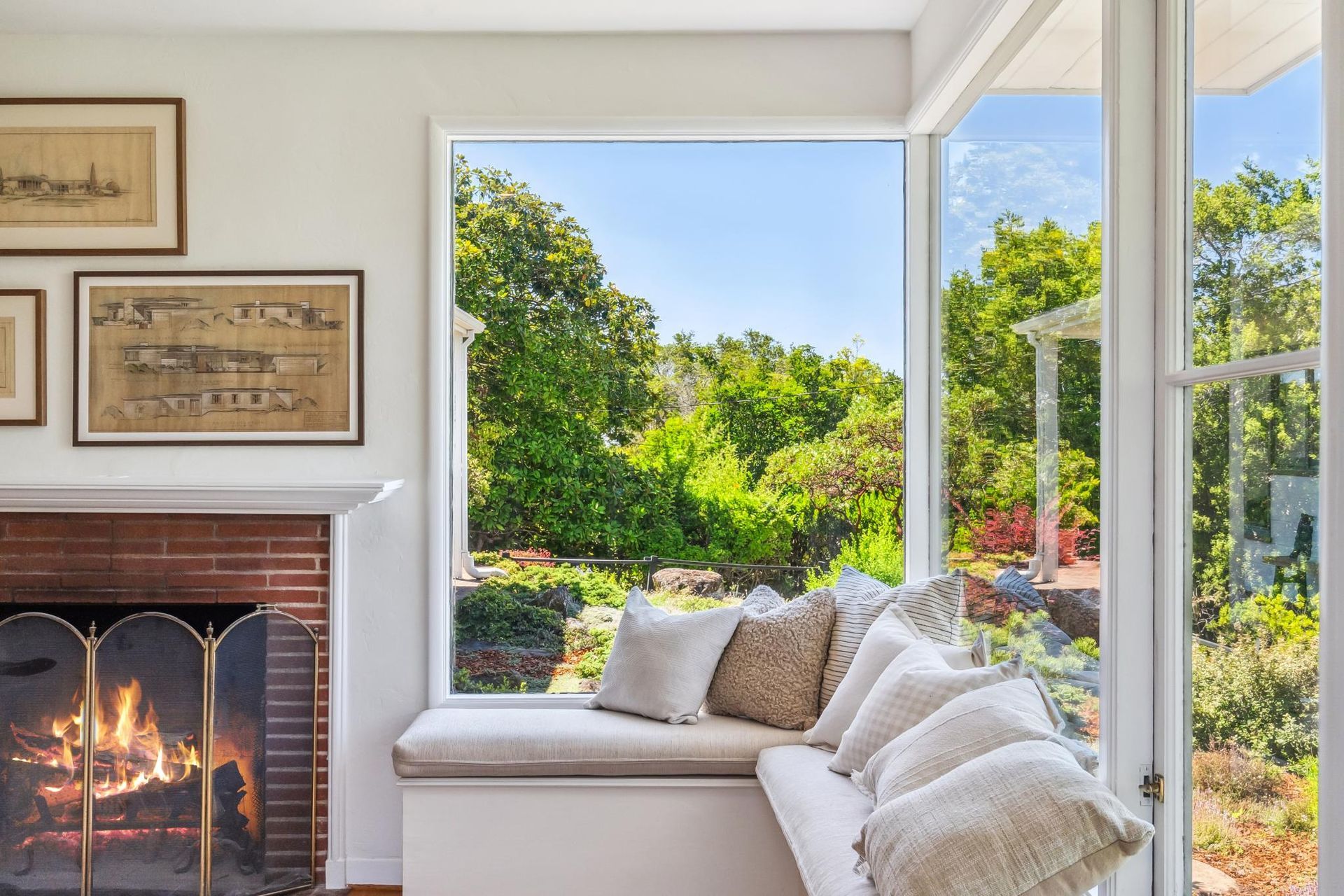 Cozy window seat with pillows overlooking a lush green garden, next to a fireplace with a burning fire.