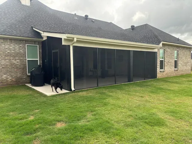 Black dog stands on a patio beside a screened porch attached to a brick house.