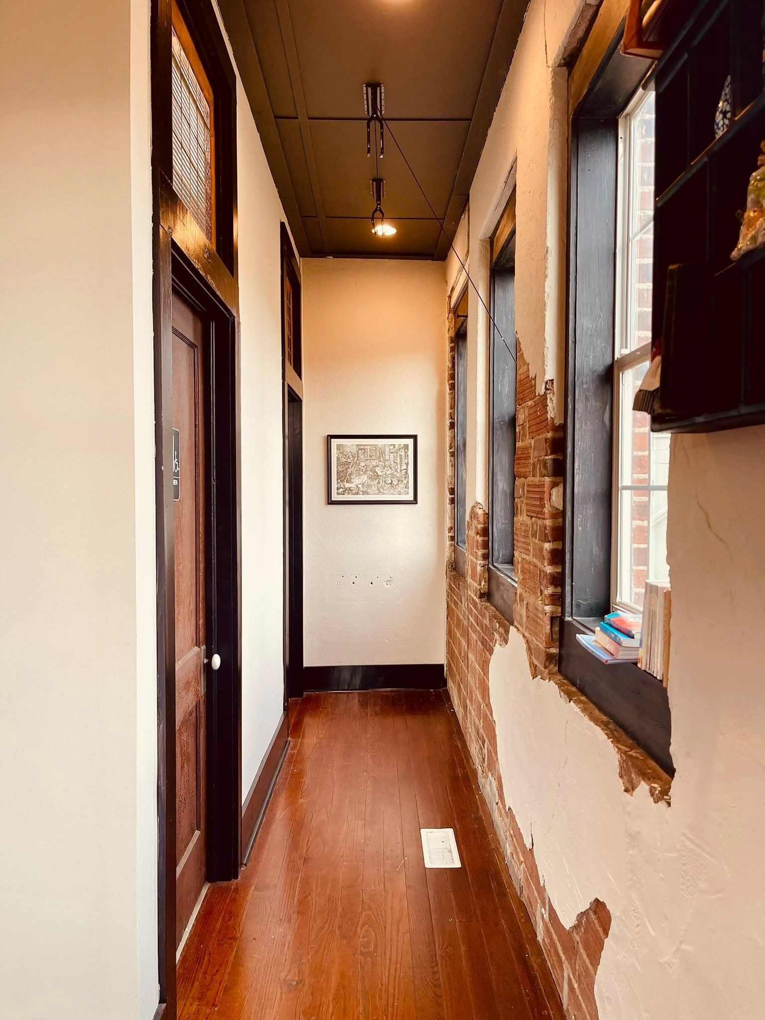 A narrow hallway with hardwood floors, dark ceiling, a door on the left, and windows with exposed brick walls on the right.