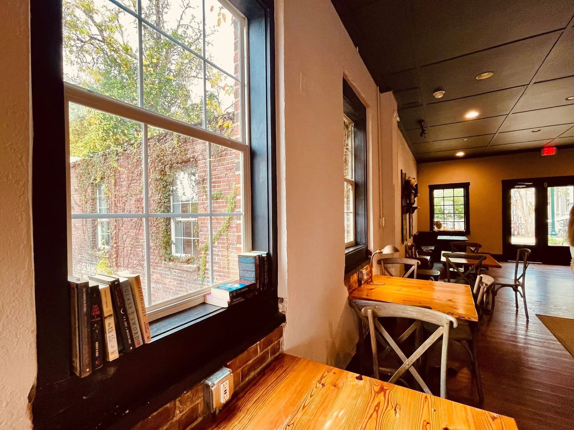 A warm, sunlit cafe interior with wooden tables, chairs, and books lined up along a window looking out onto brick walls.