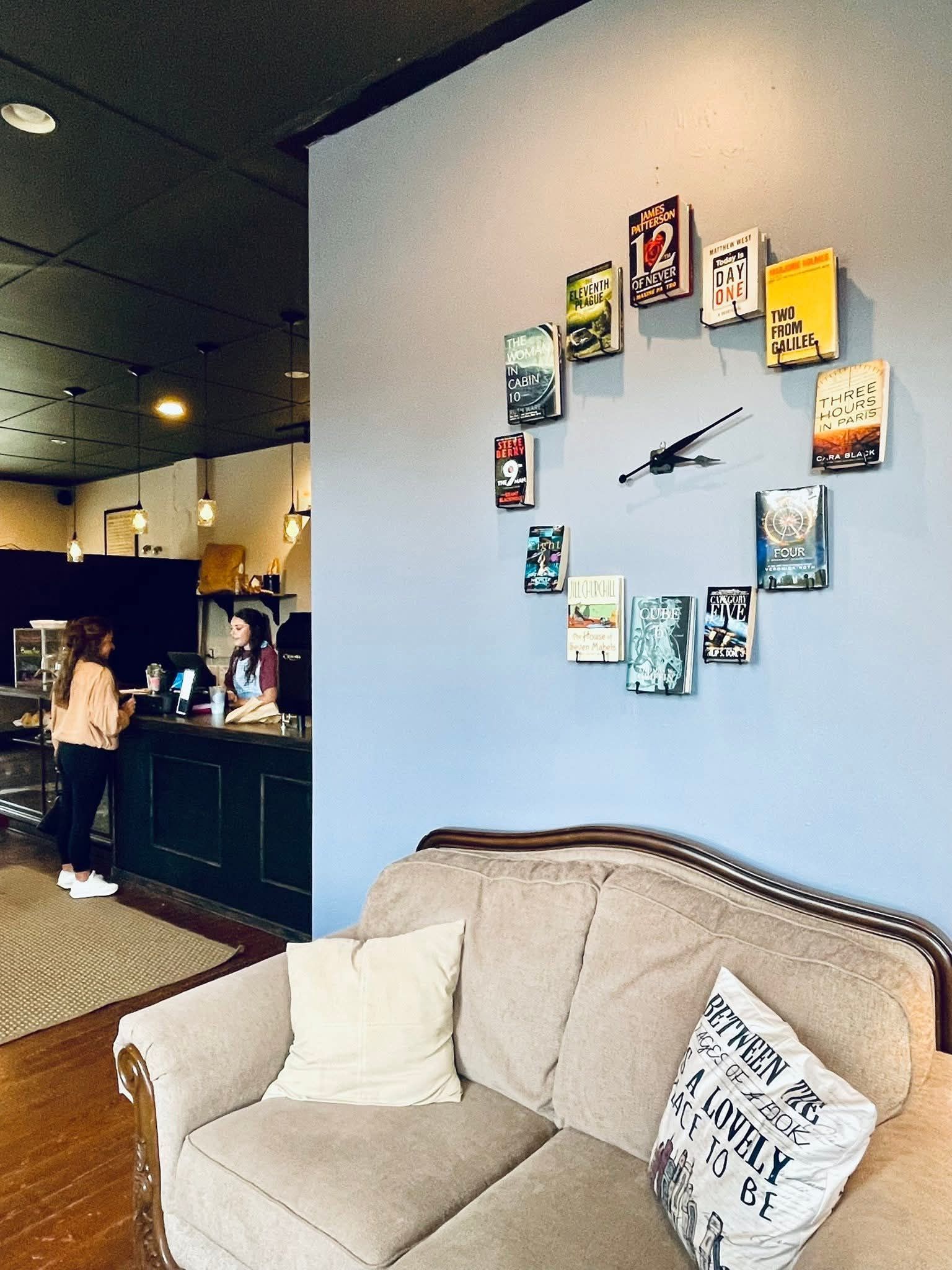 A clock on a blue wall is made of books arranged in a circle above a beige sofa inside a cafe with a service counter.
