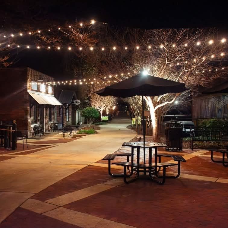 Outdoor plaza at night with a table and umbrella lit by string lights hanging between trees and buildings.