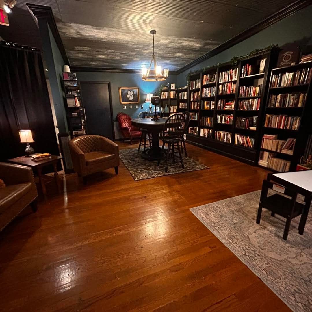 A dimly lit, wood-floored study features tall bookshelves, leather armchairs, and a round table under a chandelier.
