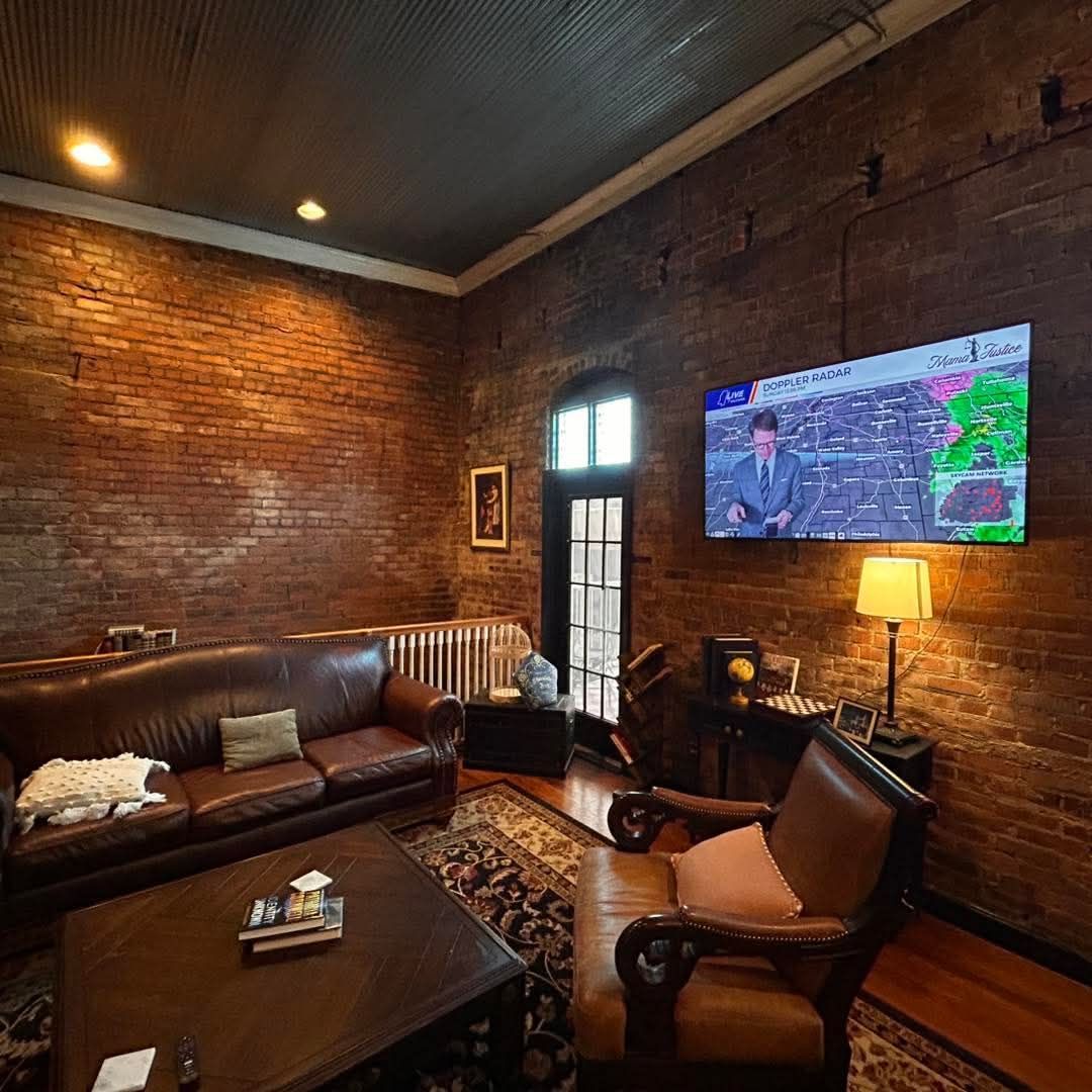 A rustic living room with exposed brick walls, a brown leather sofa, a matching armchair, and a wall-mounted television.