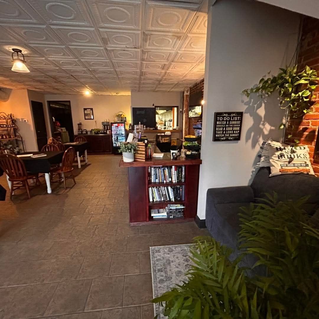 A cozy, rustic cafe interior with a bookshelf counter, tables with chairs, a tile ceiling, and a seating area with plants.