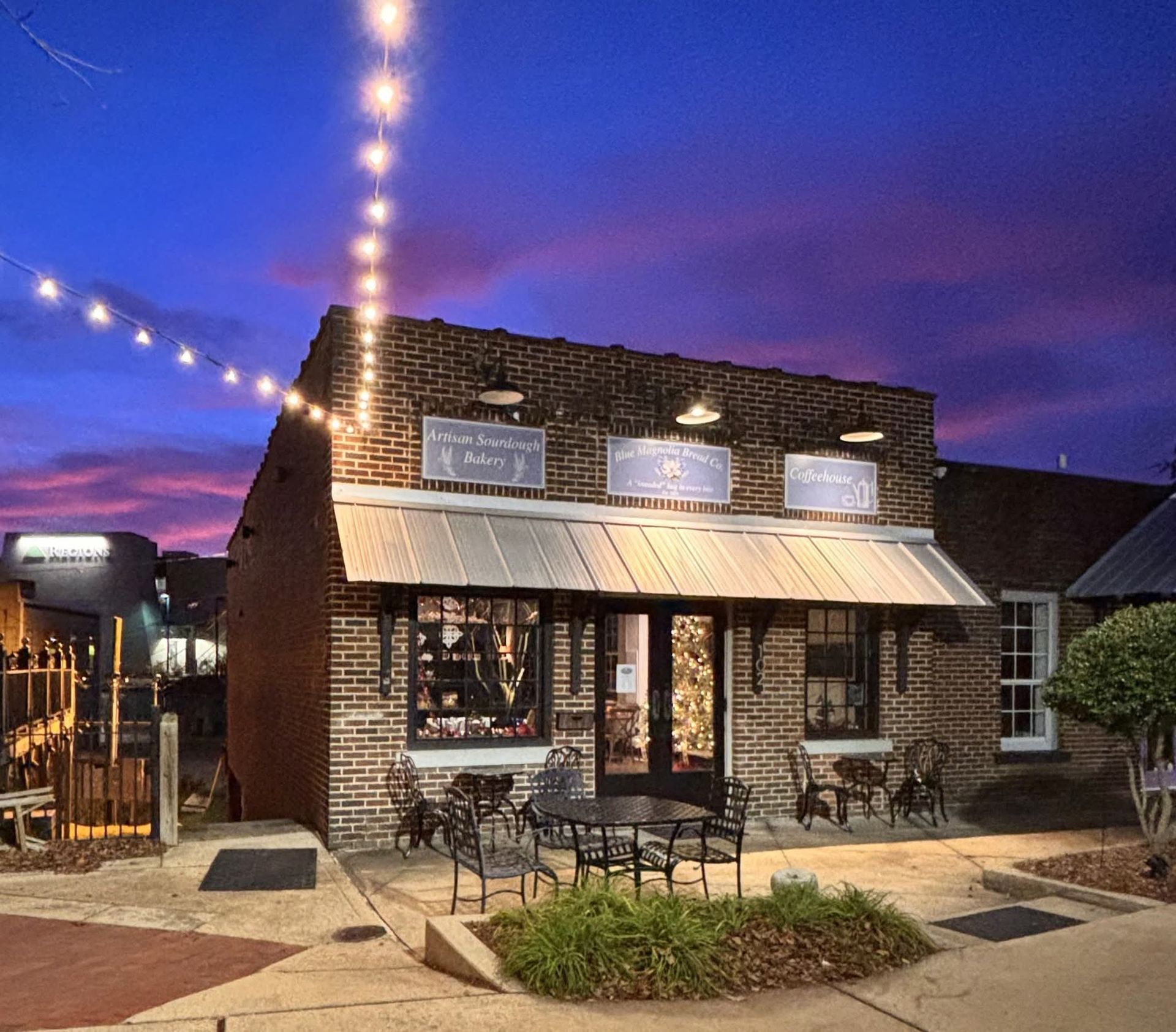 A brick commercial building with a metal awning and outdoor seating under string lights during a purple sunset.