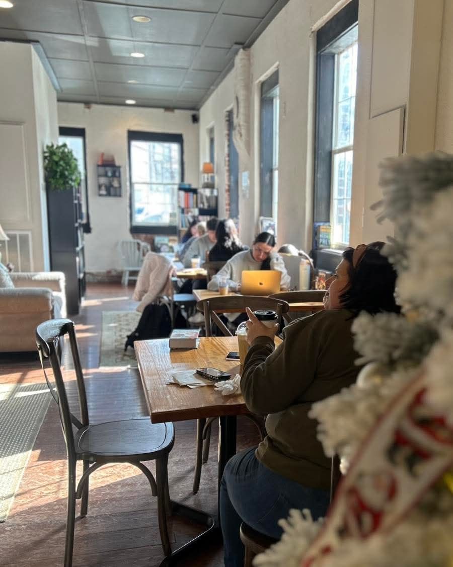 People work on laptops at wooden tables in a sunlit, narrow cafe with large windows and a festive decor in the foreground.