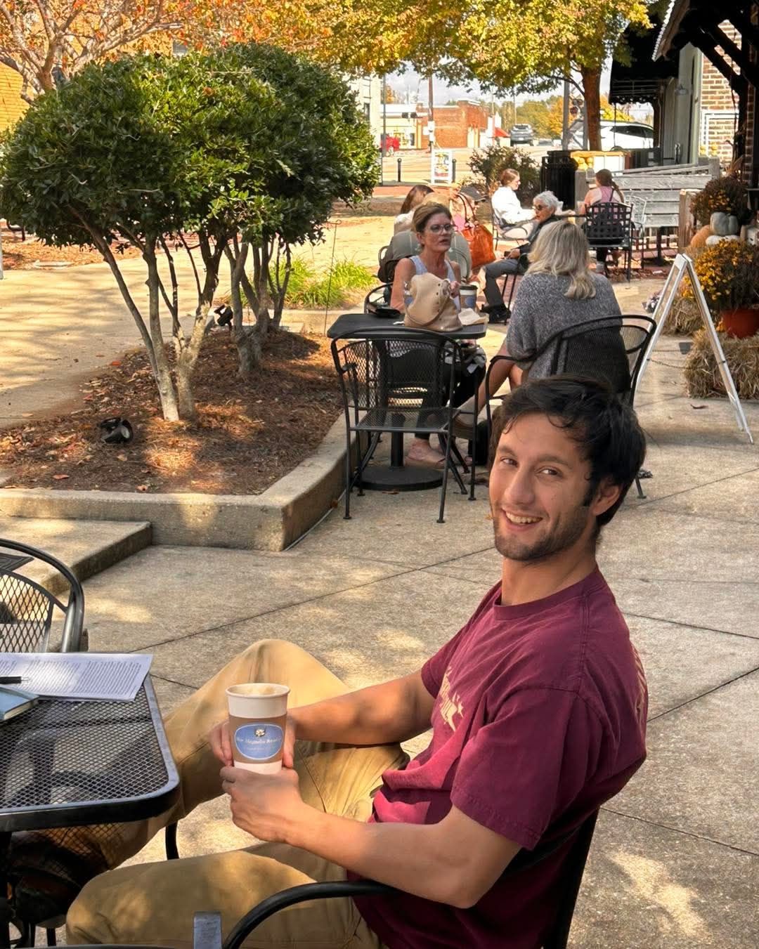 A smiling person in a maroon t-shirt sits at an outdoor cafe table holding a coffee cup, with blurred patrons behind them.